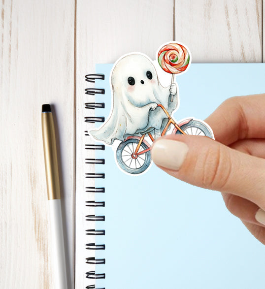 Sticker of a ghost riding a bike with a lollipop held by a hand, placed on a notebook.