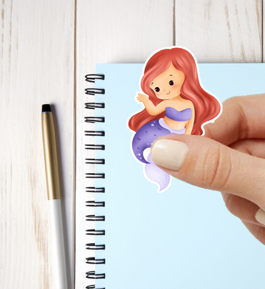Hand holding a mermaid sticker above a notebook with a pen on a light wooden surface
