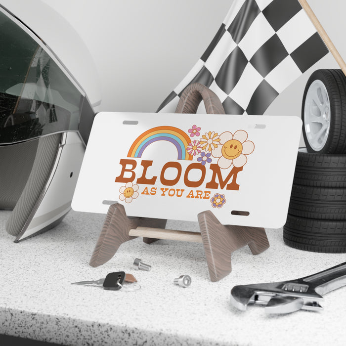 Decorative license plate with 'Bloom as you are' text and rainbow design on a stand, surrounded by car parts.